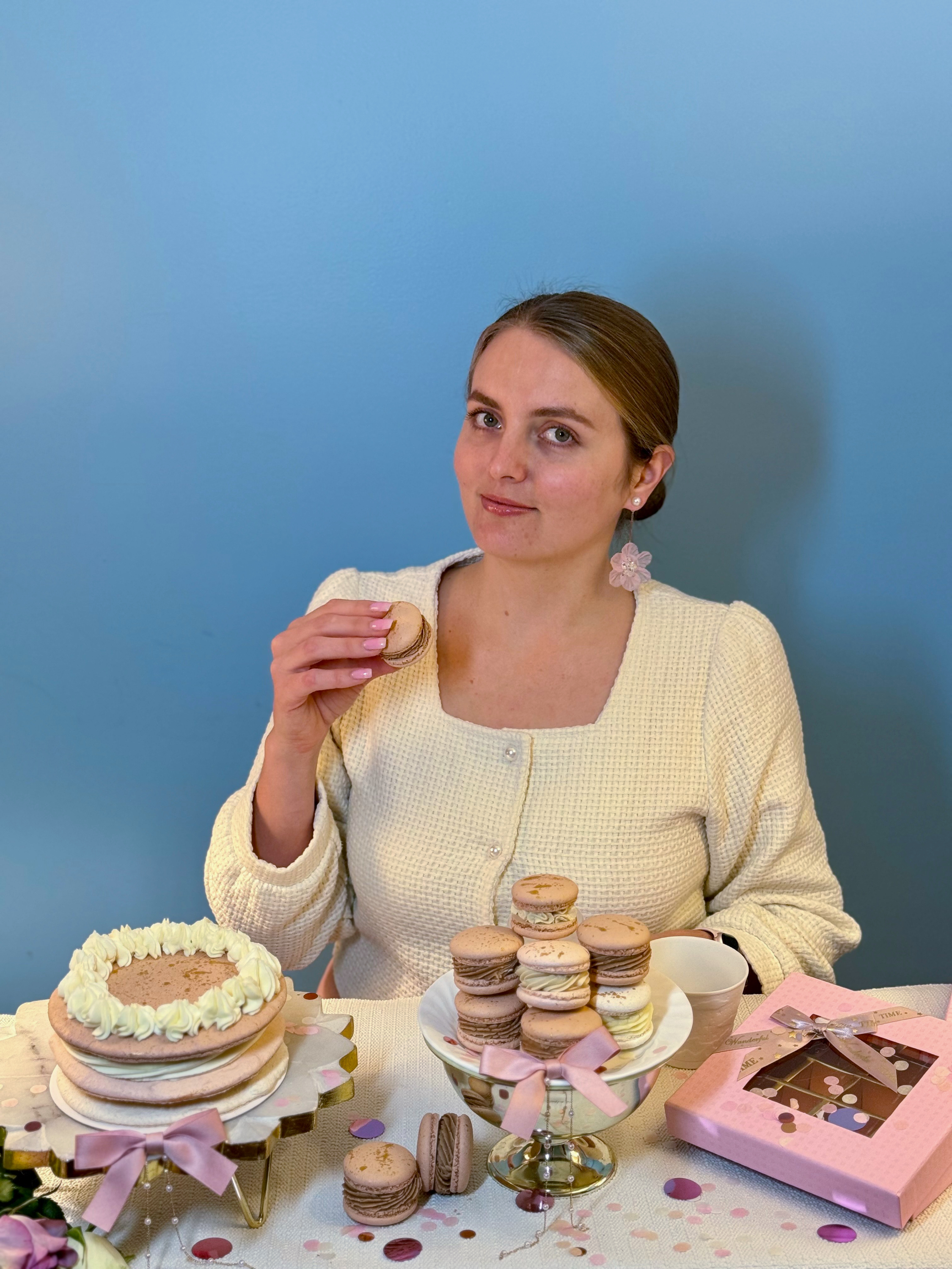 Anastasiia holding a macaron at a table set with a decorated cake, a stand of macarons and a pink gift box