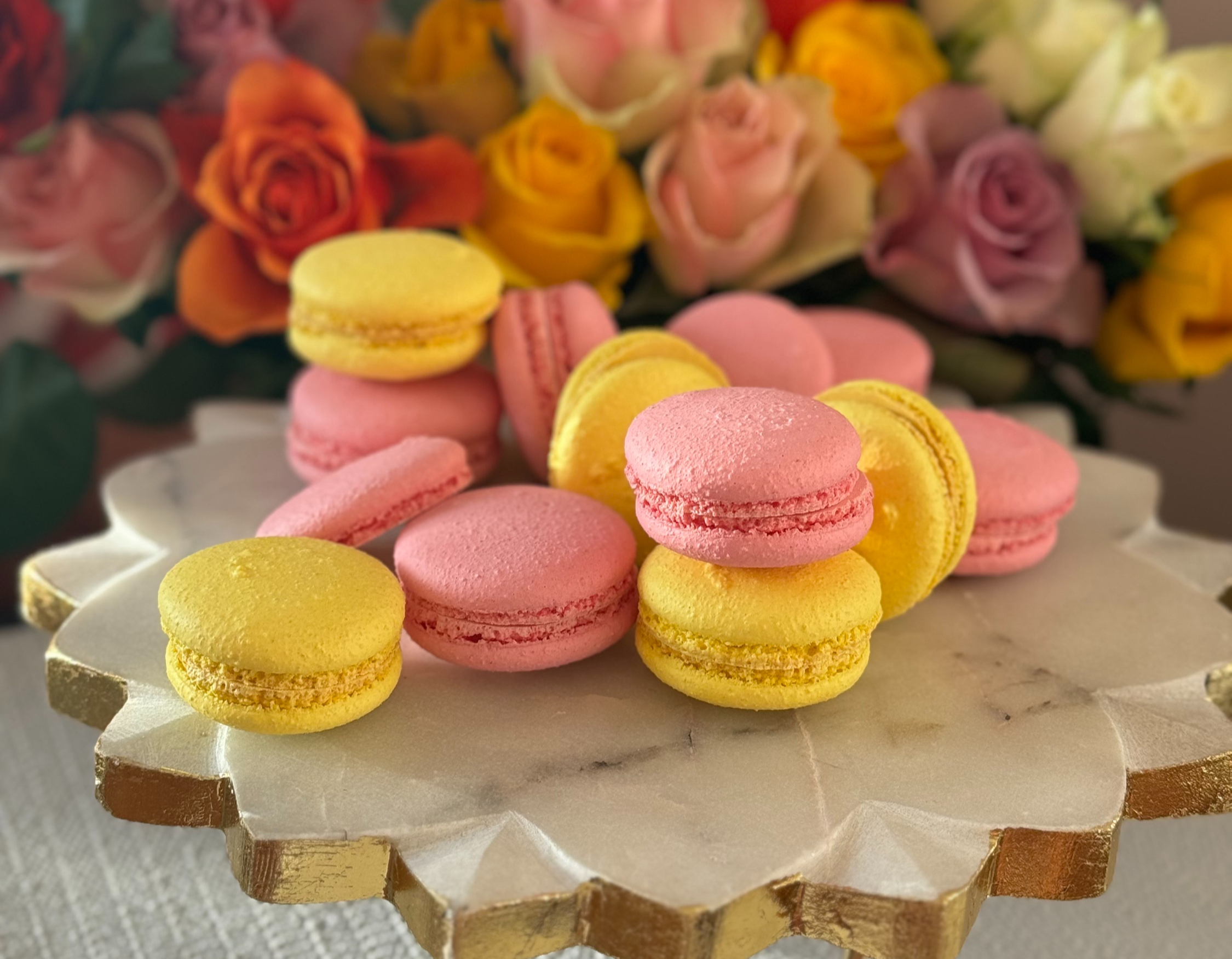 Pink and yellow macarons on a marble and gold cake stand in front of a bouquet of roses